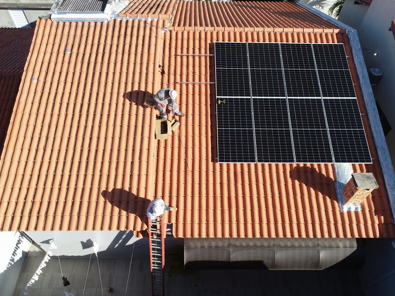 Dois técnicos de capacete instalando placas solares num telhado, vistos de drone
